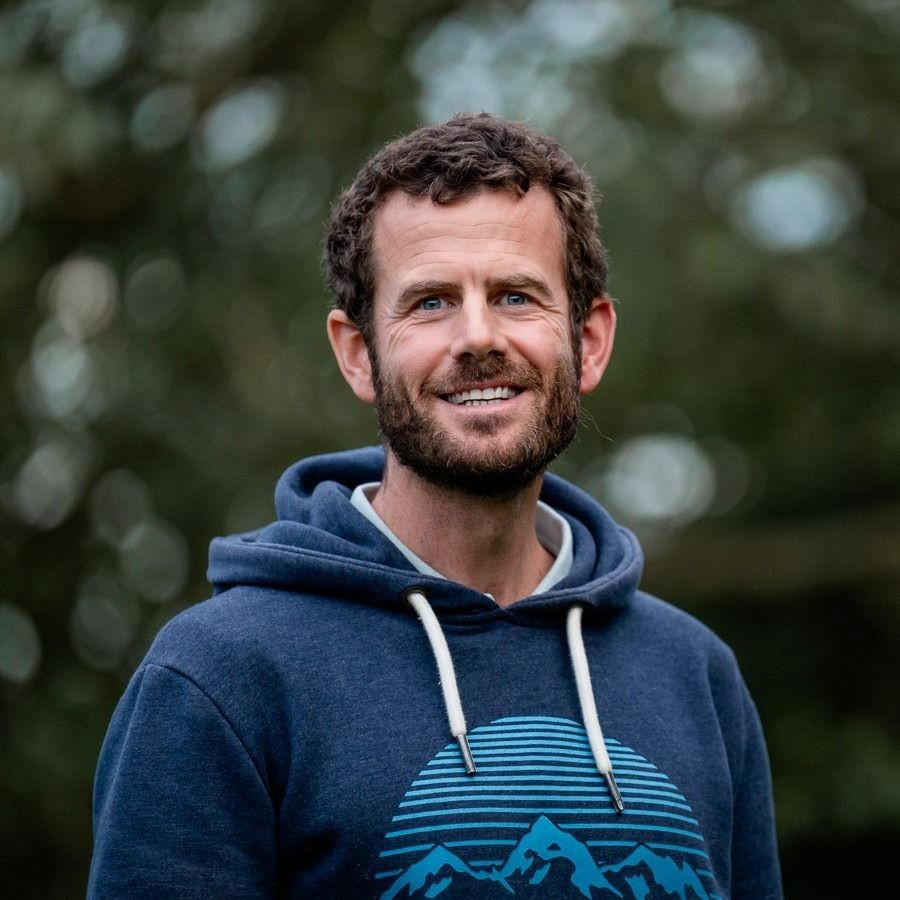 Person in a blue hoodie with mountain design standing outdoors with blurred greenery in the background.