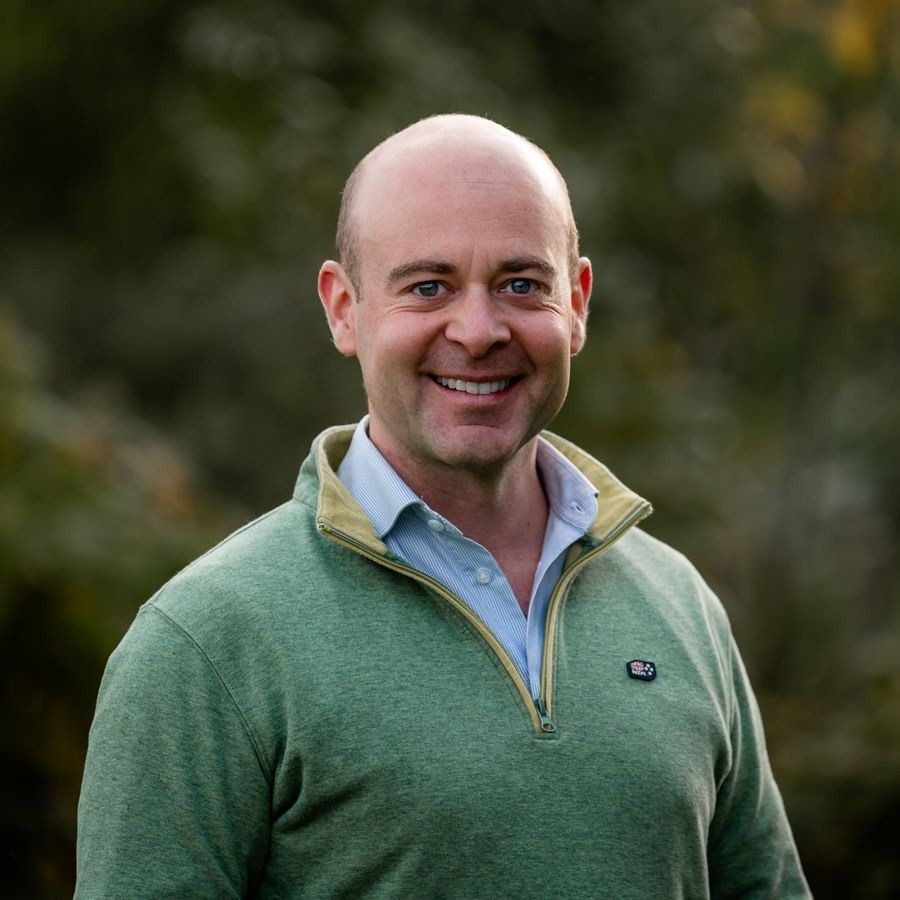 Smiling man in a green quarter-zip sweater, standing outdoors with a blurred natural background.