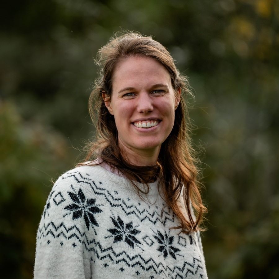 Woman smiling outdoors wearing a patterned sweater in front of blurred greenery.