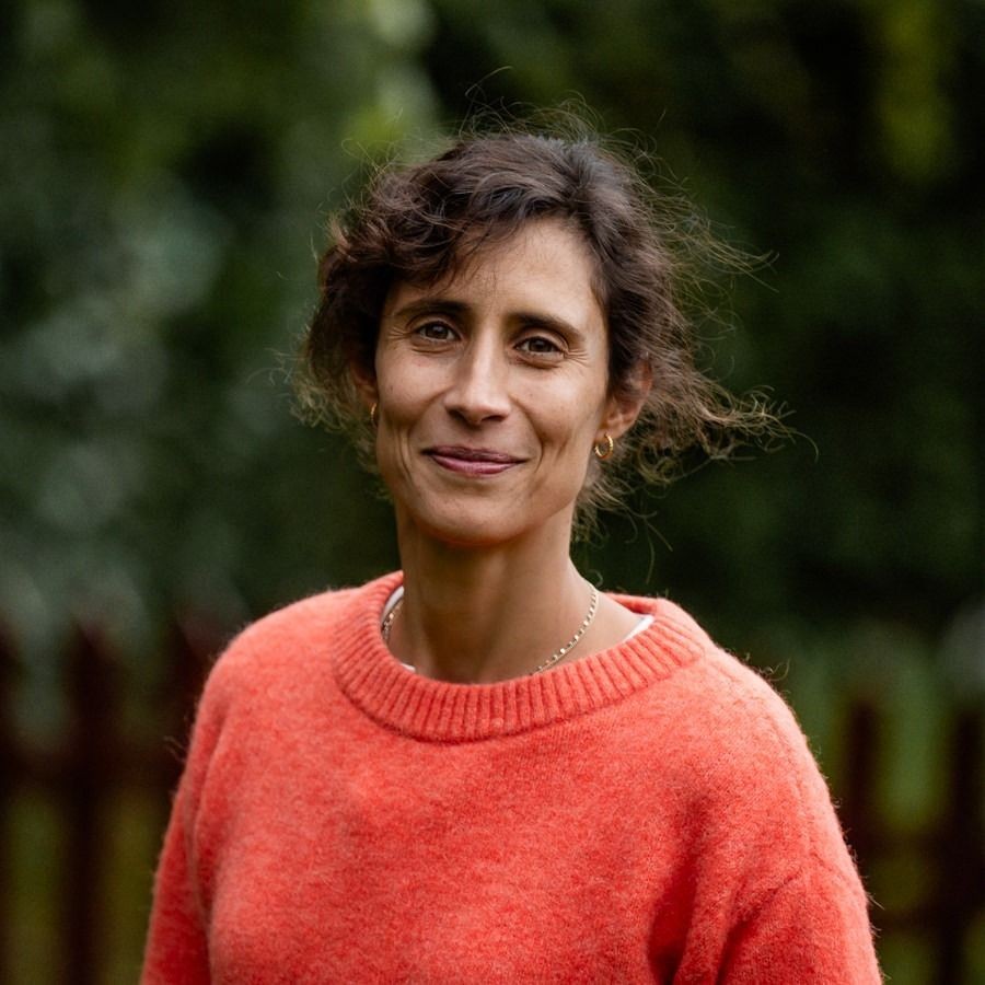 Woman smiling outdoors in a garden, wearing an orange sweater with a blurred green background.